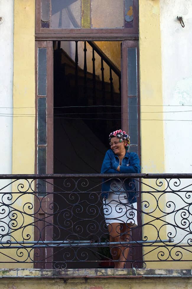 Colorful woman with curlers on her head standing on a balcony in front of an old building.